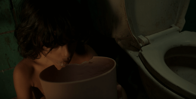A young child crouches beside a toilet and bites chunks out of a plastic garbage bin.
